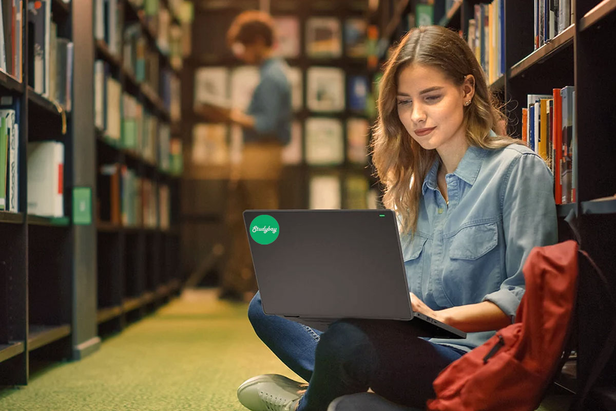 Uma garota senta-se no chão de uma biblioteca e escreve TCC on-line.